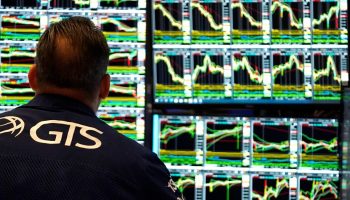 The back of the head of a stock trader is seen in front of screens that show graphs of stock movements.