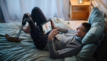 A young man with short brown hair wears a grey sweatshirt and blue pajama pants. He has a prosthetic leg. He is lying down in bed and has a tablet in his hands, resting on his waist. He is speaking to a doctor.