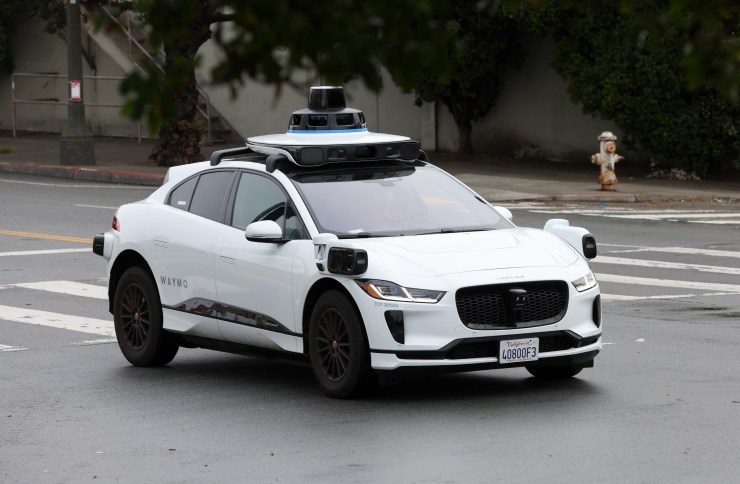 A white driverless car made by the company Waymo drives down a street in San Francisco