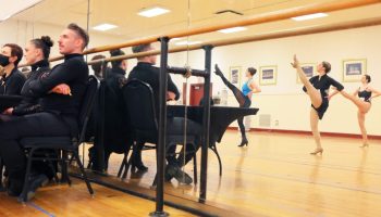 Choreographers in all black, seated at a folding table, watch dancers perform. The dancers are reflected in a ballet mirror.