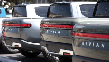A row of gray Rivian electric pickup trucks sit in a parking lot at a Rivian Service Center.