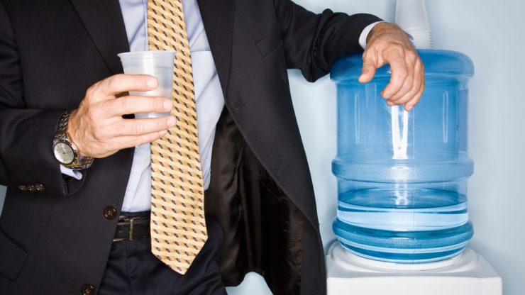 A man in a suit and tie holds a cup of water in his hand and leans on an office watercooler.