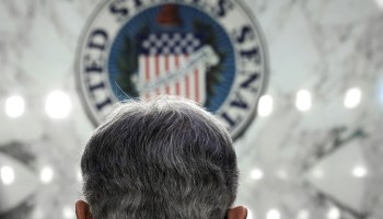 The back of Jerome Powell's head — salt and pepper hair cut short around the ears and neck — in front of a United States Senate seal.