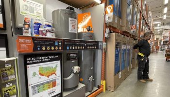 Rows of gas powered water heaters are displayed at a Home Depot store with a man standing with a tool belt on looking at shelf.