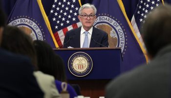 Federal Reserve Board Chairman Jerome Powell, a man with gray hair, glasses, a blue suit, white shirt and blue tie, stands at a podium with the Federal Reserve emblem and American and Federal Reserve flags. He is speaking to several rows of reporters at a press conference.