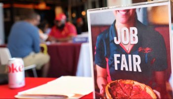 A placard at a job fair reads "Job Fair." The text is on a photo of a smiling waitress holding a pizza. The placard is on a table with a red table cloth, and people are seated at another table in the background.