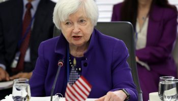 Secretary of the Treasury Janet Yellen wears a purple suit. She is sitting down speaking and a small American flag is seen situated at the table in front of her.