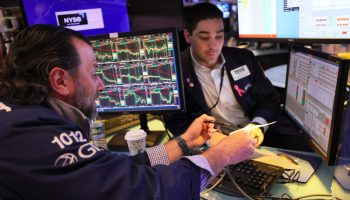 A New York Stock Exchange stock trader points toward a screen with stock movements on it. Another stock broker is nearby.