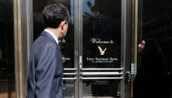 A man in a suit and a woman in a pant suit walk past a First Republic Bank branch in Manhattan. First Republic's logo is displayed on its front glass double doors.