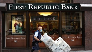 A bald, masked worker pushes a cart of white boxes as he walks past the exterior of a First Republic Bank.