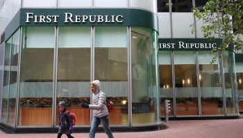 An older woman in a gray jacket and a young child in a blue jacket, both with their hoods up, walk past a First Republic Bank. It is a glass building with a green banner that reads "First Republic."