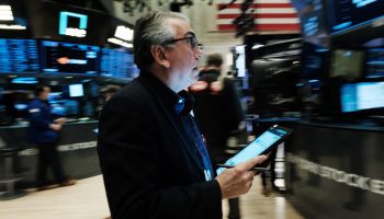 An older man with gray hair, a black blazer and black-framed glasses looks upward with his mouth open, slightly surprised. He's holding a tablet on the floor of the New York Stock Exchange. He is surrounded by screens displaying various stock movements.