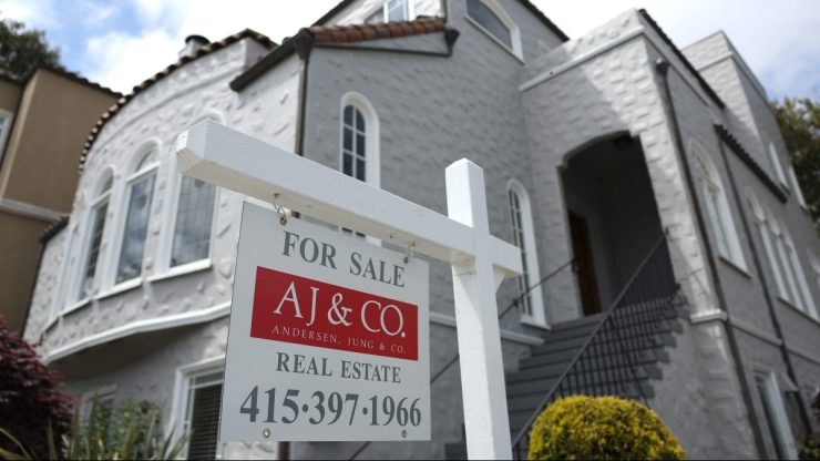 A for sale sign is posted in a multi-story gray stucco house with red terra cotta roof shingles.