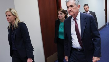 Federal Reserve Chairman Jerome Powell, with salt and pepper hair, wearing a white shirt, dark suit and maroon tie, walks down a hallway with three other people.