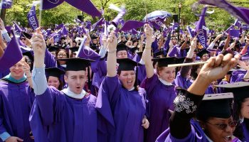 A crowd of college graduates in deep purple robes and black motarboards wave pennants and cheer.