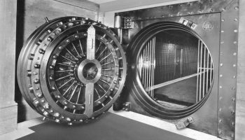 A black and white image shows the round door to a massive vank vault.
