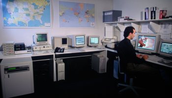 A man sits at a 1990s-era computer. Several other small, old computers are on the desk. On the walls are maps of the world and the United States, and a shelf with a few books and a box.