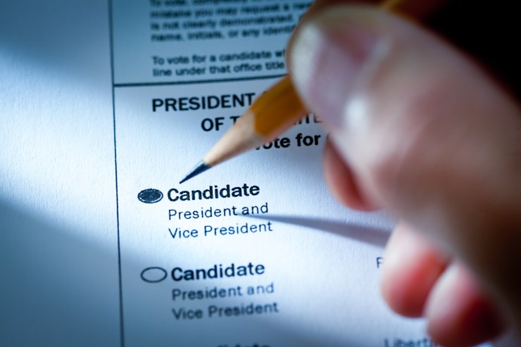 A close-up macro photograph of the United States Presidential Election Voting Ballot with a hand holding a pencil filling out the selected candidate. Photographed in dramatic lighting spotlighting the selection and the word "Candidate". Selective focus on the ballot selection of President and Vice President.