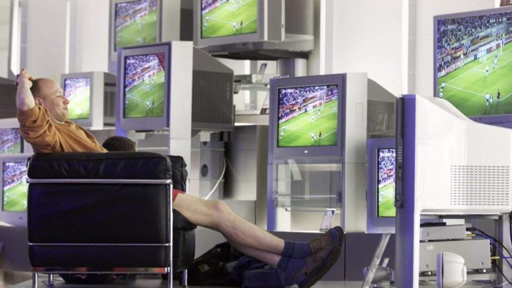 A man reclines in a black chair with hands above his head in front of several TV screens playing a sporting game.