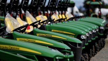 A row of new John Deere tractors, with green hoods and yellow stripes bearing the maker's name. The black steering wheels are visible, along with yellow seats covered in plastic.