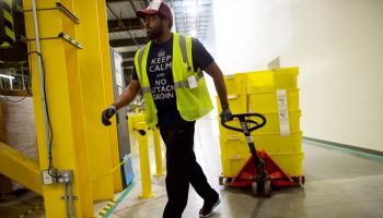 A Black man wearing a neon yellow vest and dark jeans pulls yellow containers around a warehouse.