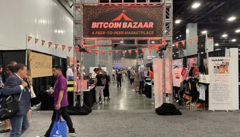 I black sign hangs with red lettering stating, "Bitcoin Bazaar" at Bitcoin 2023, a Bitcoin convention in Miami Beach. Booths are set up behind the sign and people are walking around.