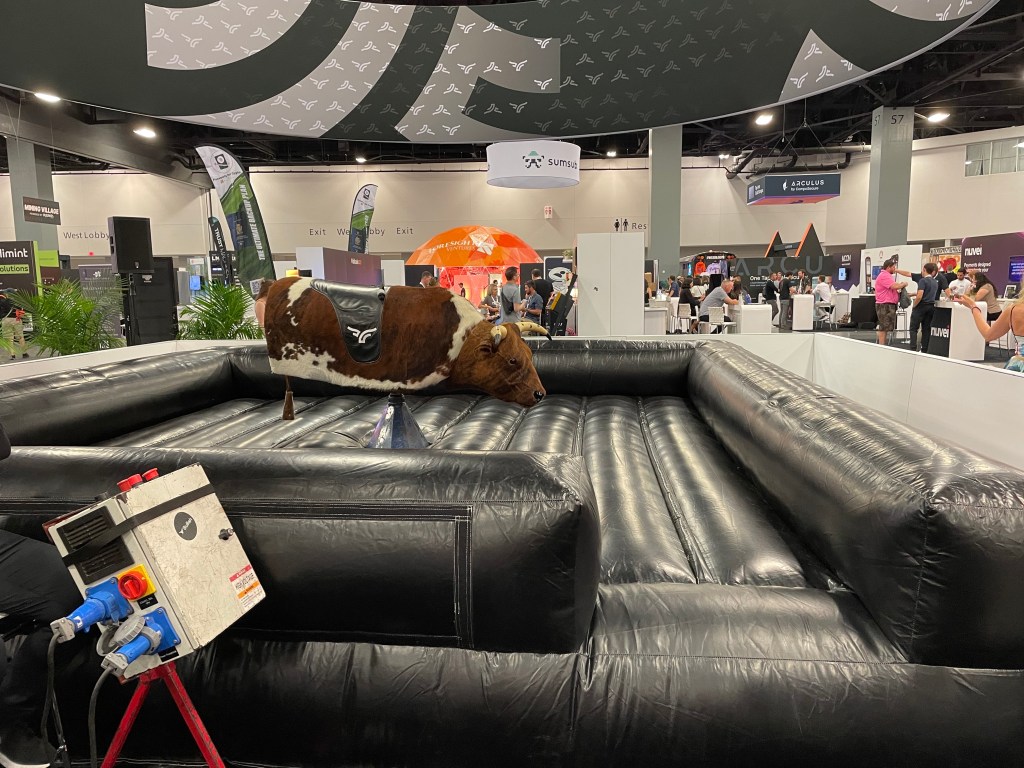A brown and white mechanical bull sits in the middle of a large, black inflatable landing pad on the floor of the Bitcoin 2023 convention hall.