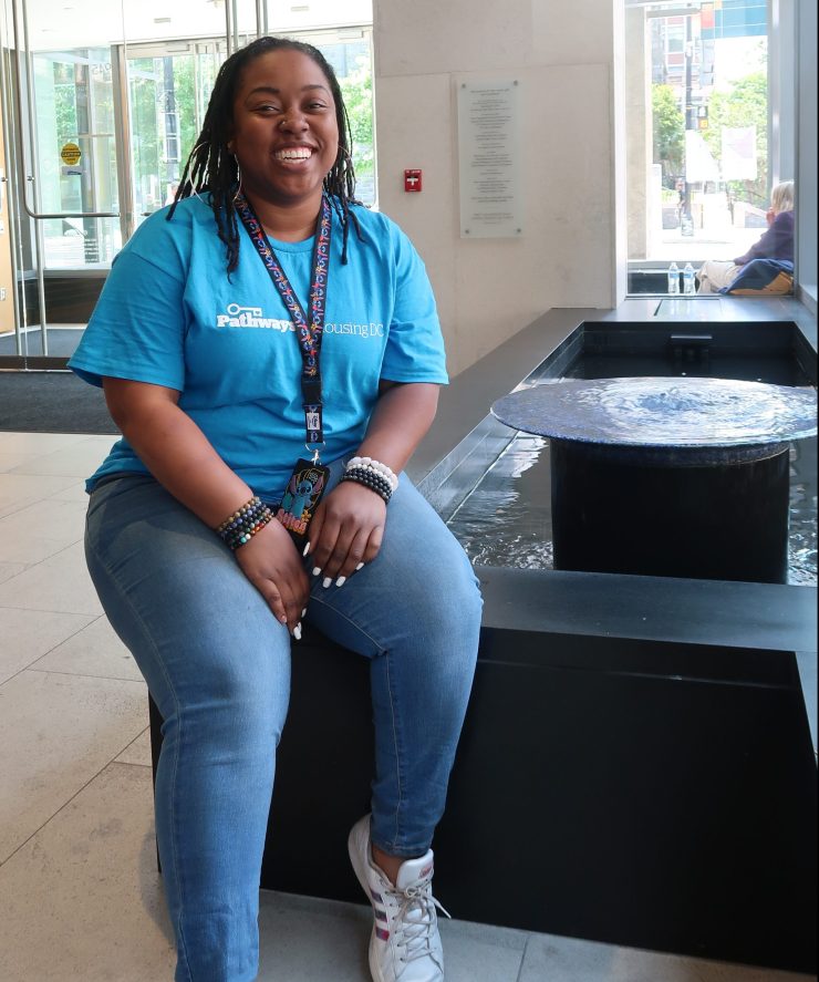 Grace Lee sits on the corner of a low indoor fountain. She is a Black woman with locks wearing jeans, white sneakers, and a blue t-shirt that says "Pathways to Housing DC". She has a big smile and black lanyard around her neck with her ID.