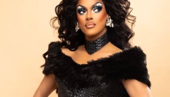 A drag queen with full makeup poses in a black, form fitting dress with a thick black necklace and big earrings.