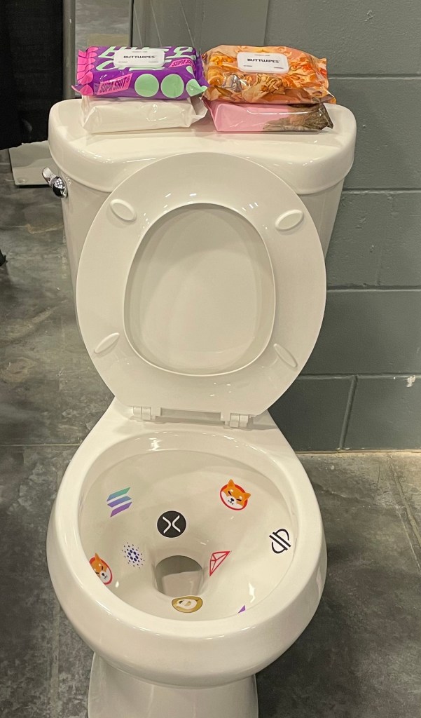 A disconnected white toilet with stickers bearing the logos of various cryptocurrencies sits on the floor of a convention hall. On top of the toilet's tank are four packages of wet wipes.