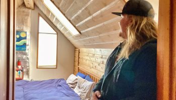 Amber Kennedy's roommate, a woman wearing a blue jacket and black baseball cap, has long blonde hair. She is standing in the doorway of an attic bedroom.