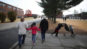 On a sidewalk, two women in hijabs, long pants and jackets hold the hands of a small girl in a bright pink jacket. All are walking away from the camera down the road. Next to them, a child pushes a smaller child in a stroller. Near them are trees and air force base buildings used for temporary housing.