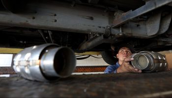 Under the body of a car, the head and hands of a car mechanic is seen. He's looking up, installing one of four new catalytic converters.