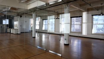 An office with glass walls, two white pillars, large windows and wood floors sits entirely empty.
