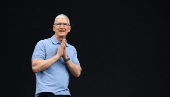 Apple CEO Tim Cook is an older man with short, white hair, dark-rimmed glasses, black pants and a light blue polo. He is speaking in front of a solid black background and has both hands clasped together as if in prayer. He is speaking.