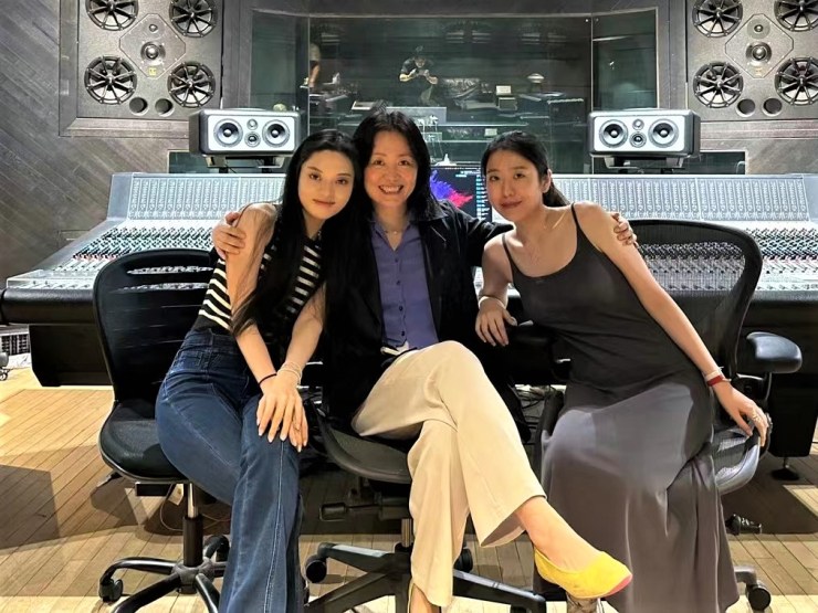Three women sit together in office chairs in front of a sound board. iQiyi Wonderworks’ Wang Huiyu sits in the middle with her arm around the other two women. She has black hair, a blue shirt, light pants, yellow shoes and a black jacket. 