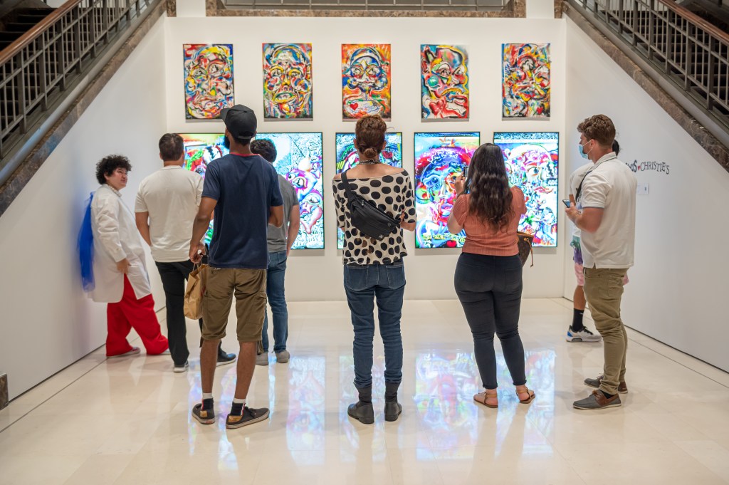 Nine people look at a wall displaying five abstract, colorful paintings above five TVs displaying NFTs similar to the art above.
