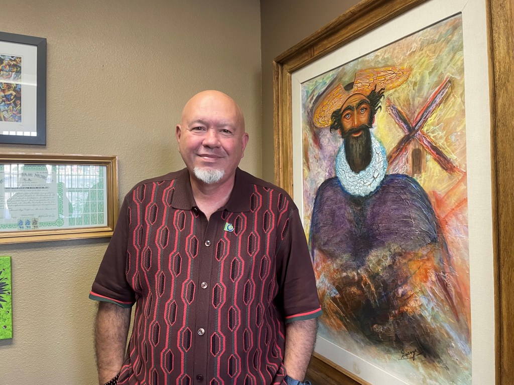 Carlos Gomez, the vice president of business development at the Latin Chamber of Commerce of Nevada, wears a red shirt and standards near a Don Quixote portrait within his office. 