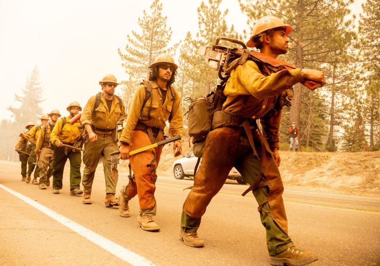 A U.S. Forest Service firefighter crew walks in line on a road in a smokey forest setting.