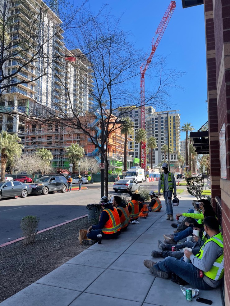 Contruction workers in neon vests sit in the shade during a lunch break in downtown Phoenix, Arizona in February, 2024. In the background are new apartments under construction. 