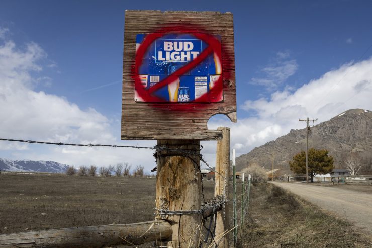 On a wooden fence post in a rural setting, part of a Bud Light package is visible with red spray paint of a slashed circle on top of it.